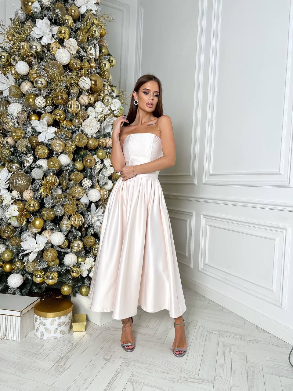 Woman in a white dress standing next to a decorated Christmas tree in a room with white walls and flooring.