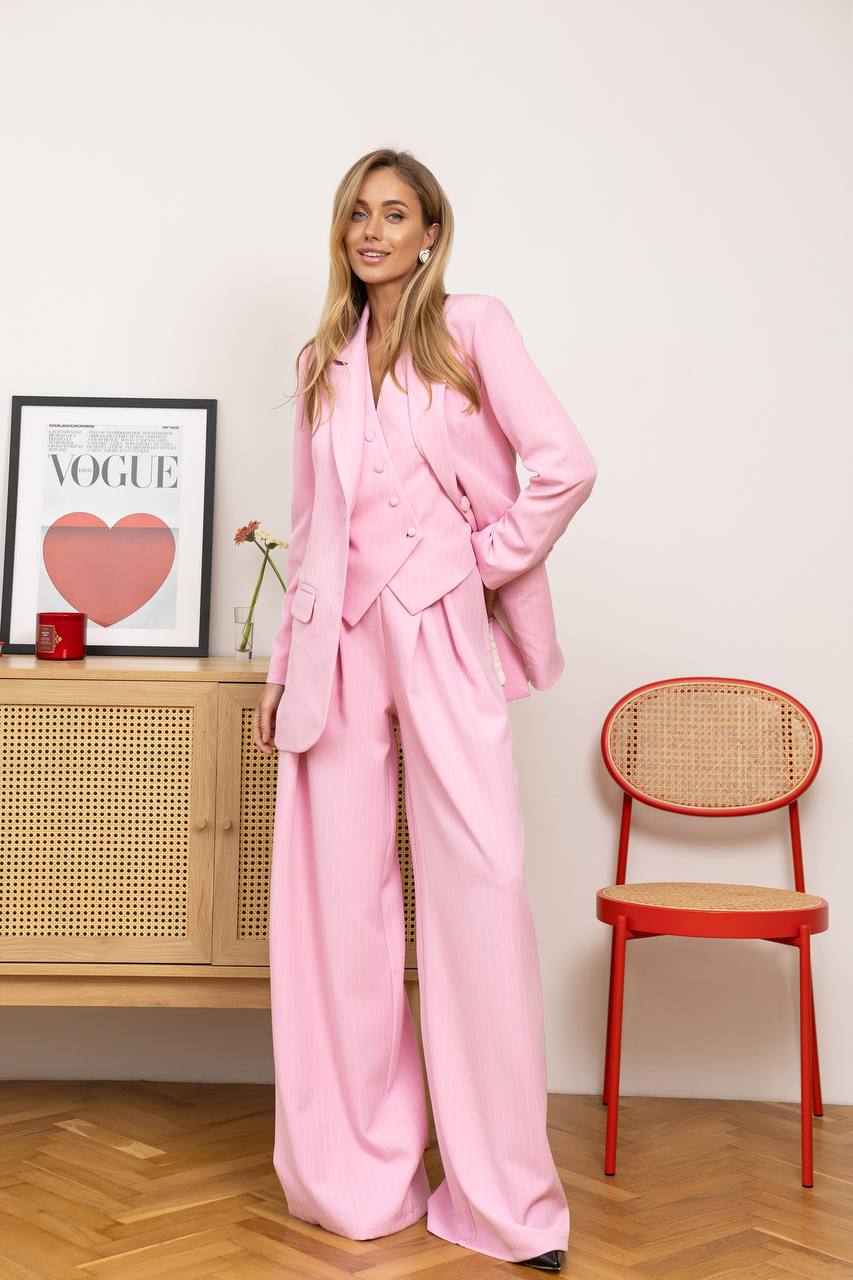 Woman in a pink suit standing in a room with a red chair and framed picture on a shelf.
