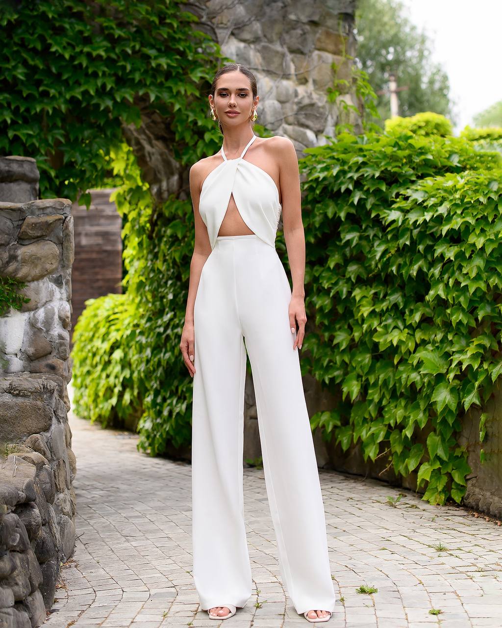 Woman in a white outfit standing in a garden with greenery and stone walls.