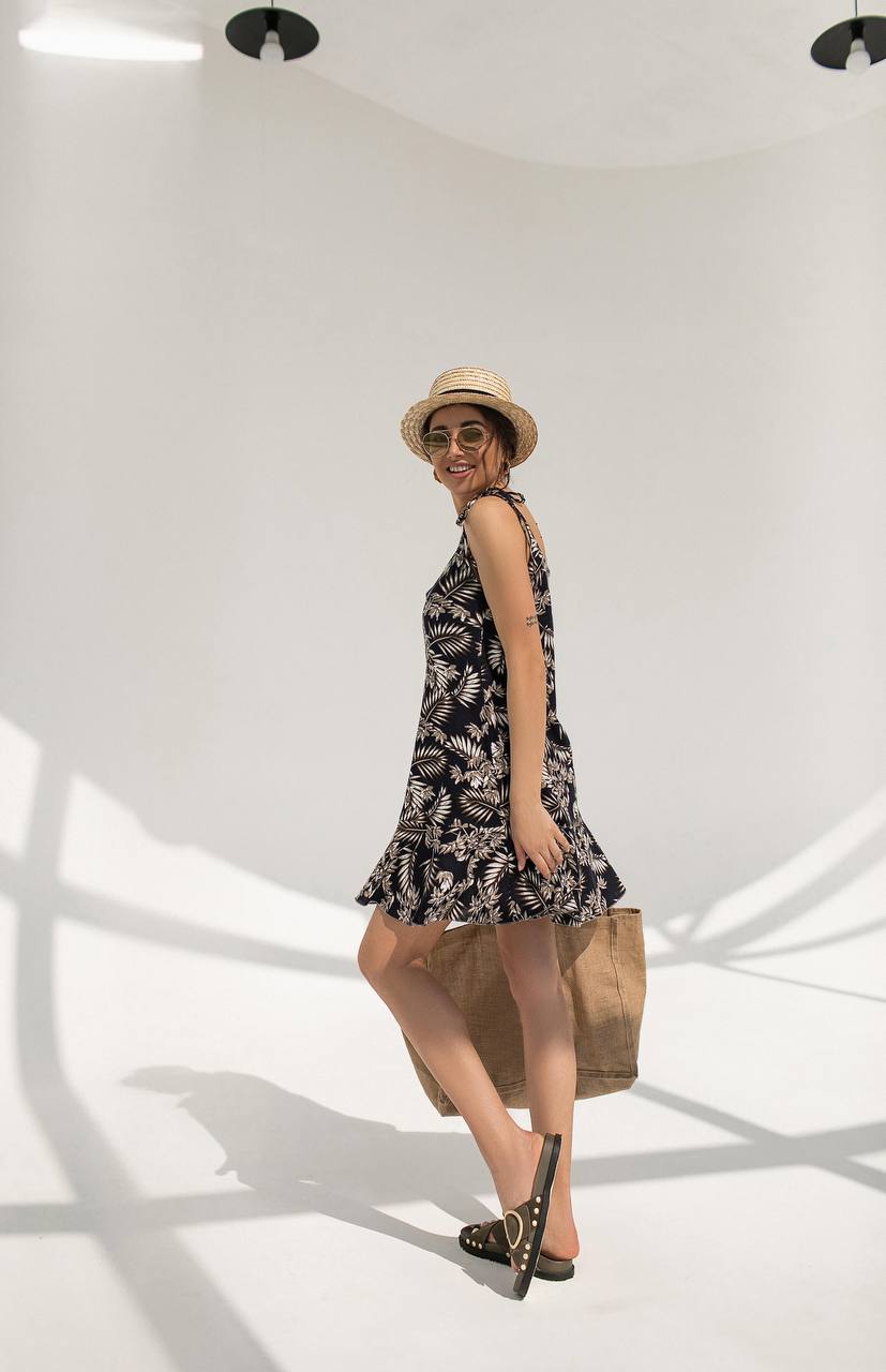 Woman in a black floral dress and straw hat standing in a well-lit room with shadows on the floor.