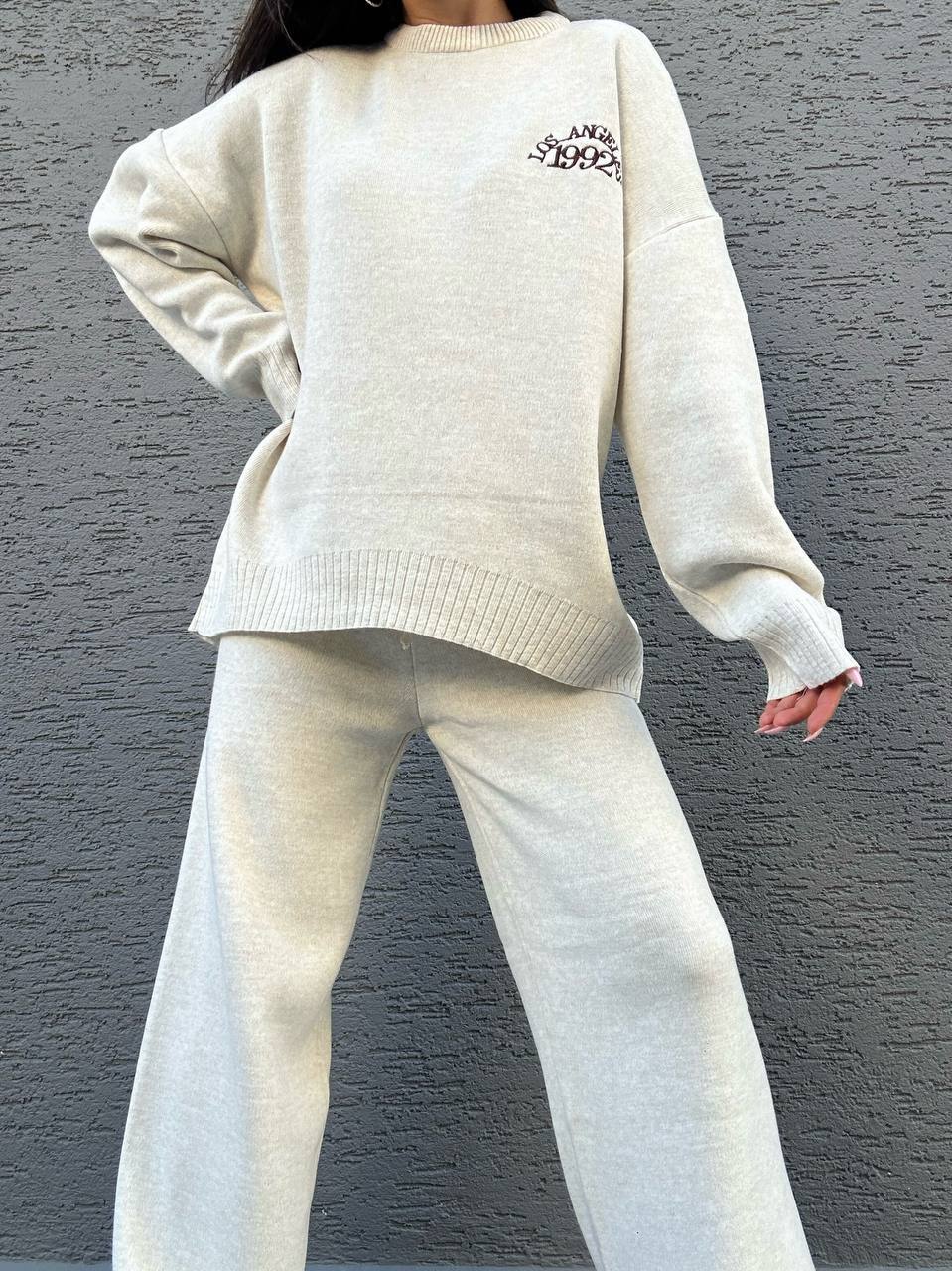 Person wearing a cream-colored sweater and pants against a gray wall