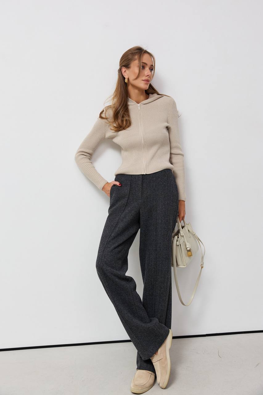 Woman wearing a beige sweater and gray pants against a white background