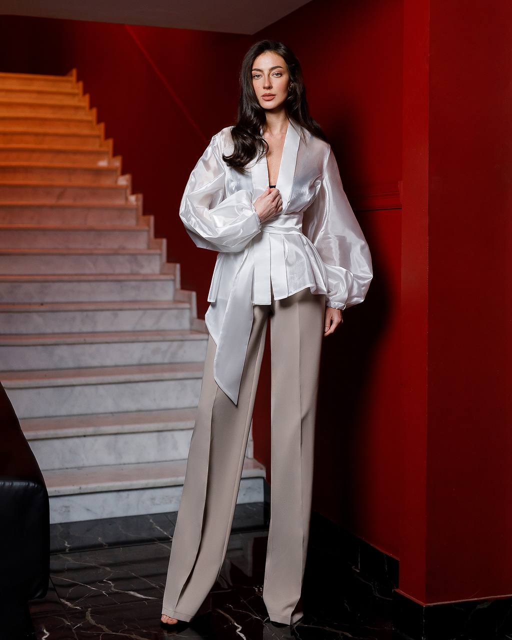 Woman in a white blouse and beige pants standing in front of a red wall with a staircase.