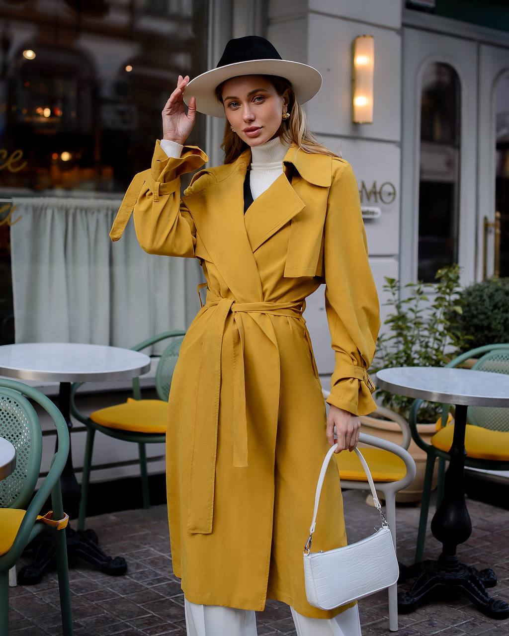 Woman in a yellow trench coat and white hat standing in an outdoor cafe setting.