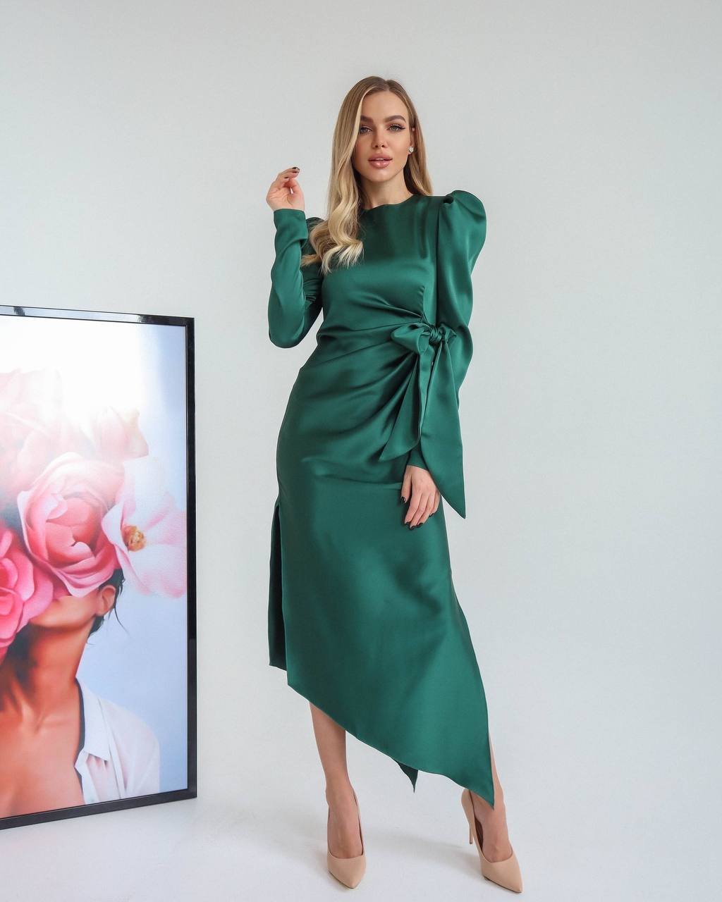 Woman wearing a green dress standing in front of a white wall with a framed picture of a woman with flowers.