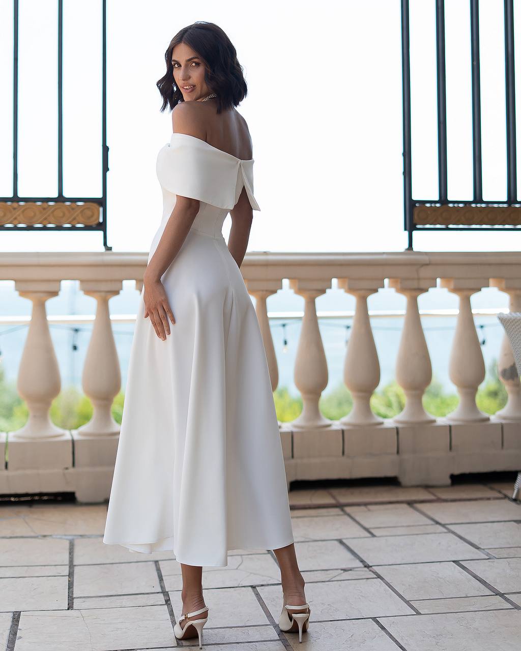Woman in a white off-shoulder dress standing on a balcony with a scenic background.