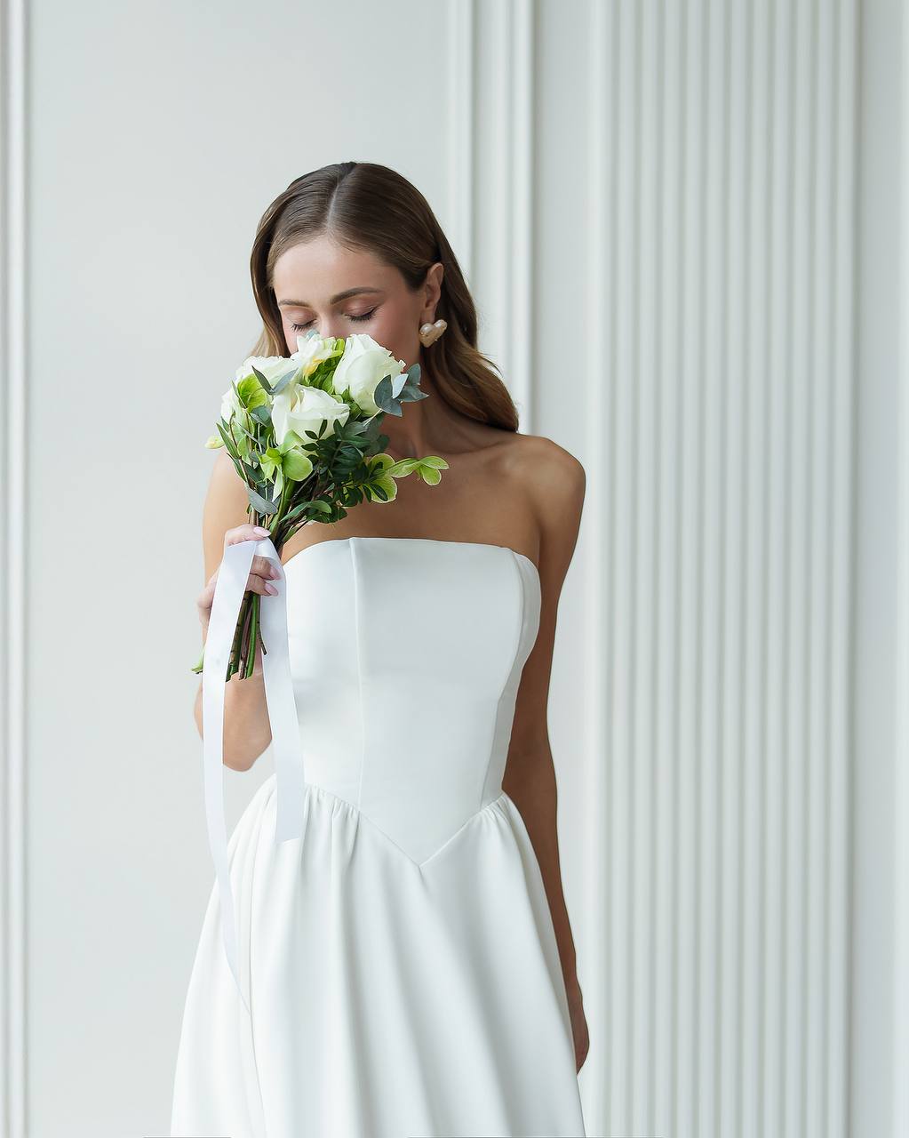 Woman in a white strapless dress holding a bouquet of flowers against a light gray background