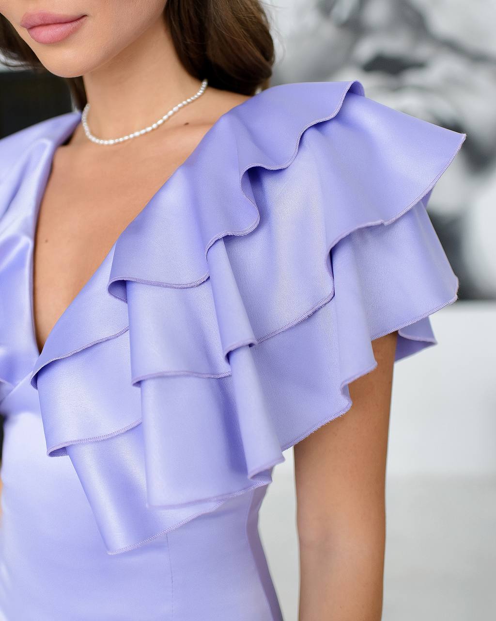 Close-up of a person wearing a light purple dress with ruffled shoulders against a neutral background.