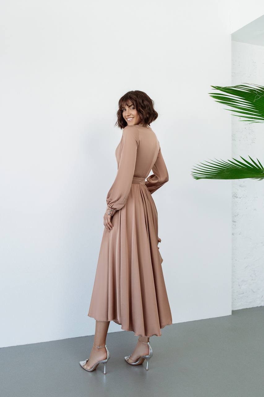 Woman in a beige dress standing against a white wall with a plant in the corner.
