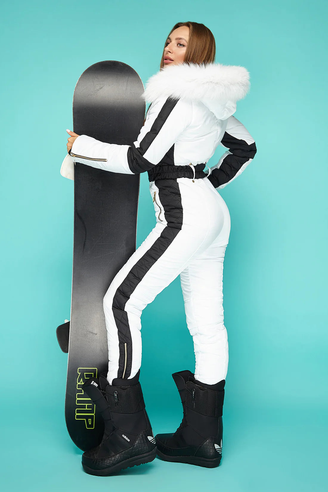 Person in a white snowsuit with black accents holding a snowboard against a teal background