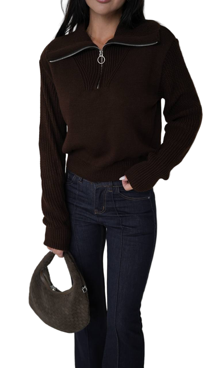 Person wearing a brown sweater and dark jeans holding a brown handbag on a white background