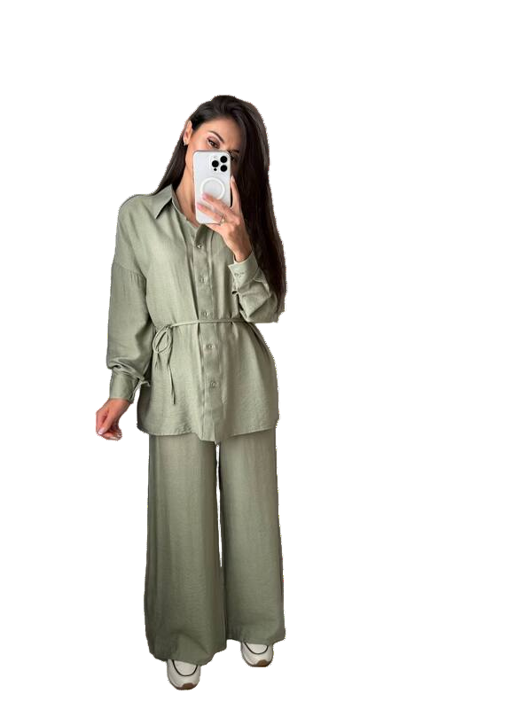 Person wearing a green outfit with wide-leg pants, holding a phone, and standing next to a small handbag on a white background.