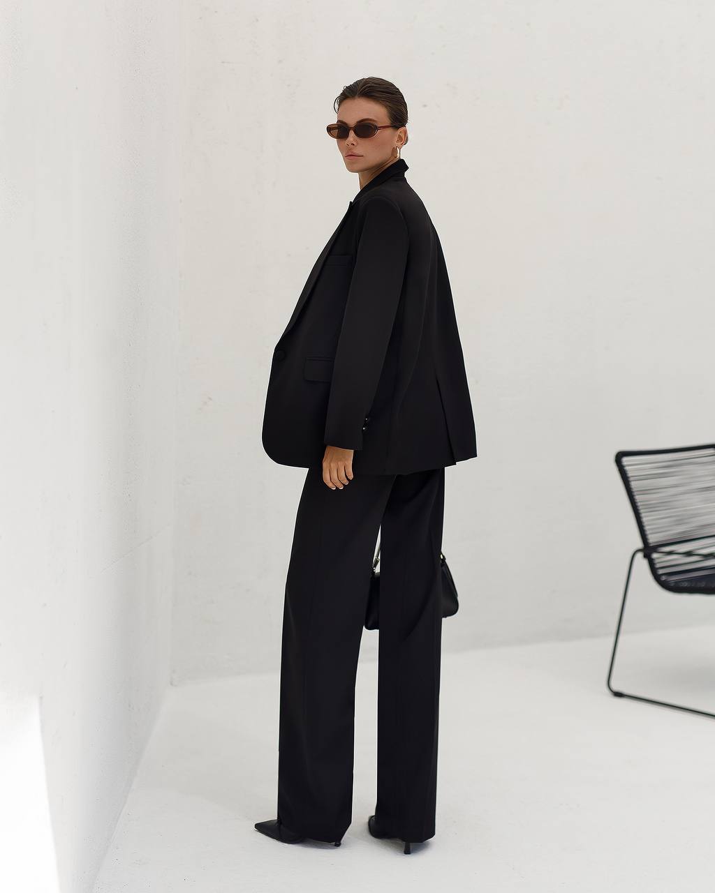 Person wearing a black suit standing against a white background with a chair visible.