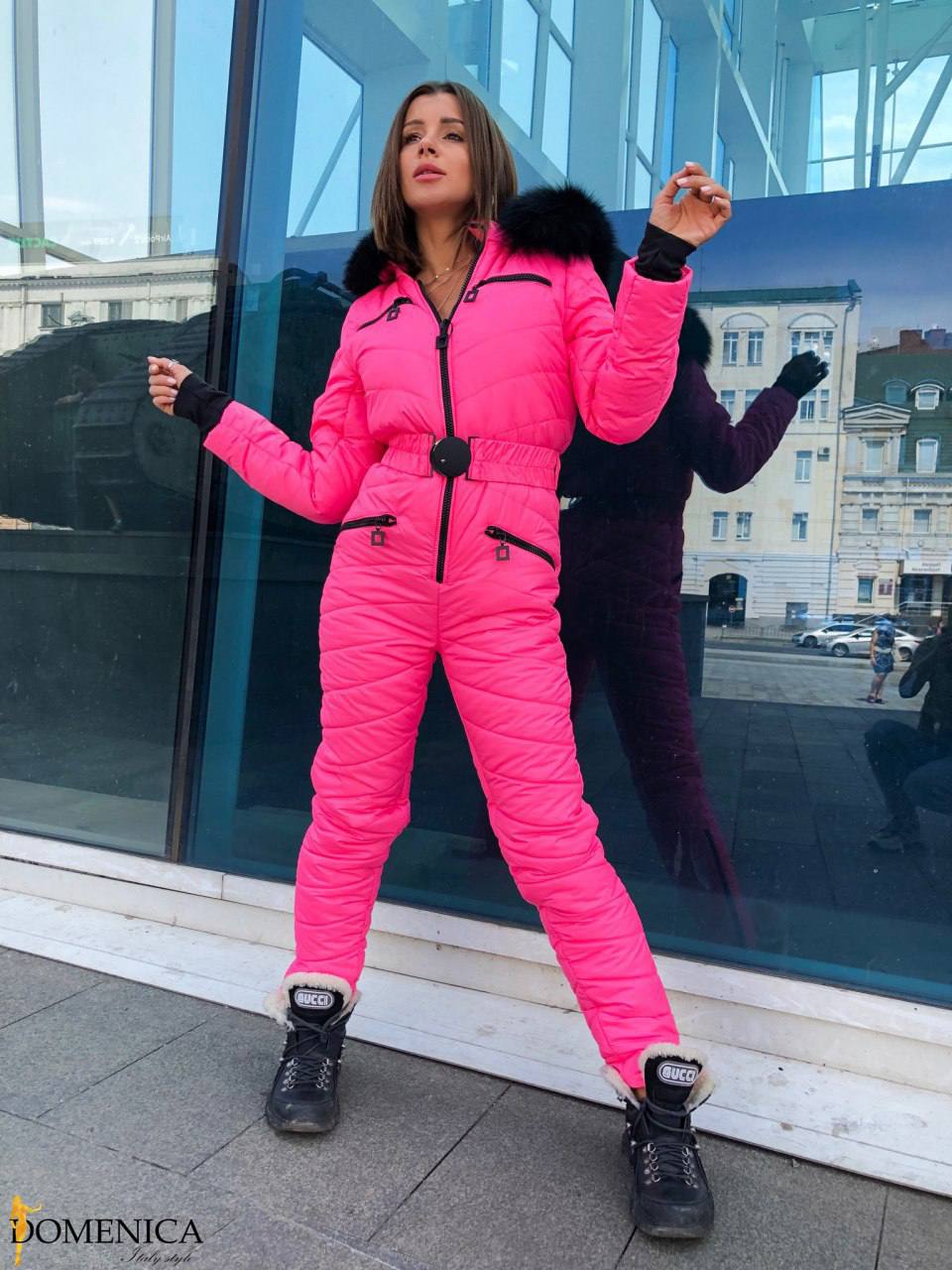 Person wearing a bright pink puffer jumpsuit with black accents, standing in front of a reflective glass surface.