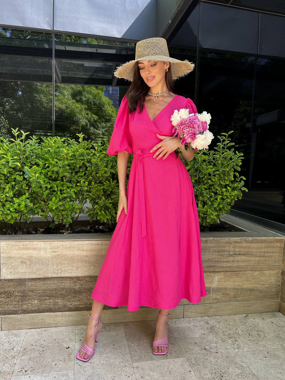Woman in a bright pink dress and hat holding flowers outdoors.
