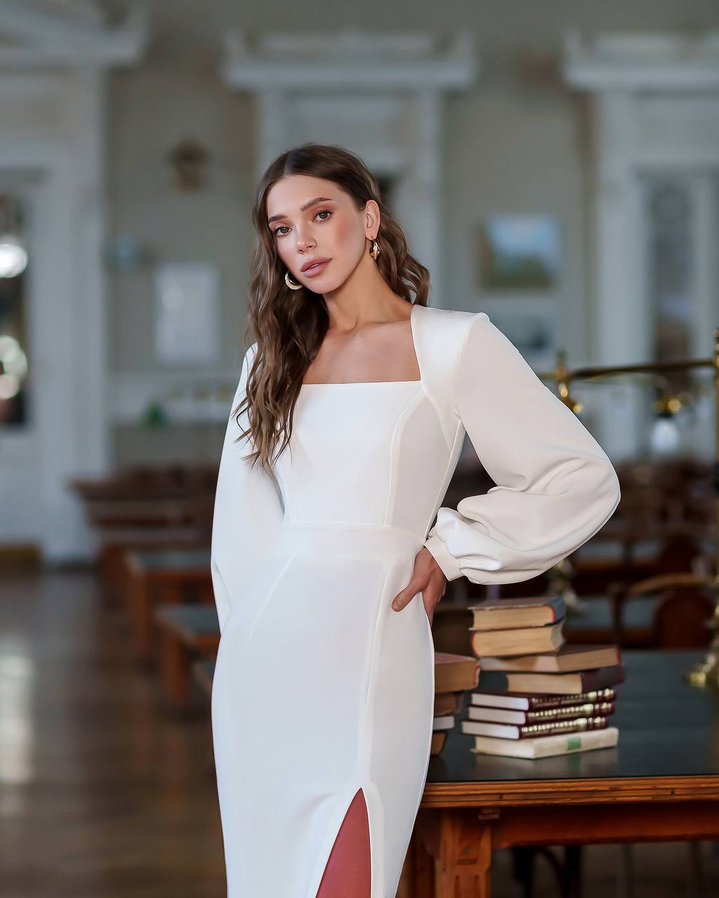 Woman in a white dress standing in an elegant room with books on a table.