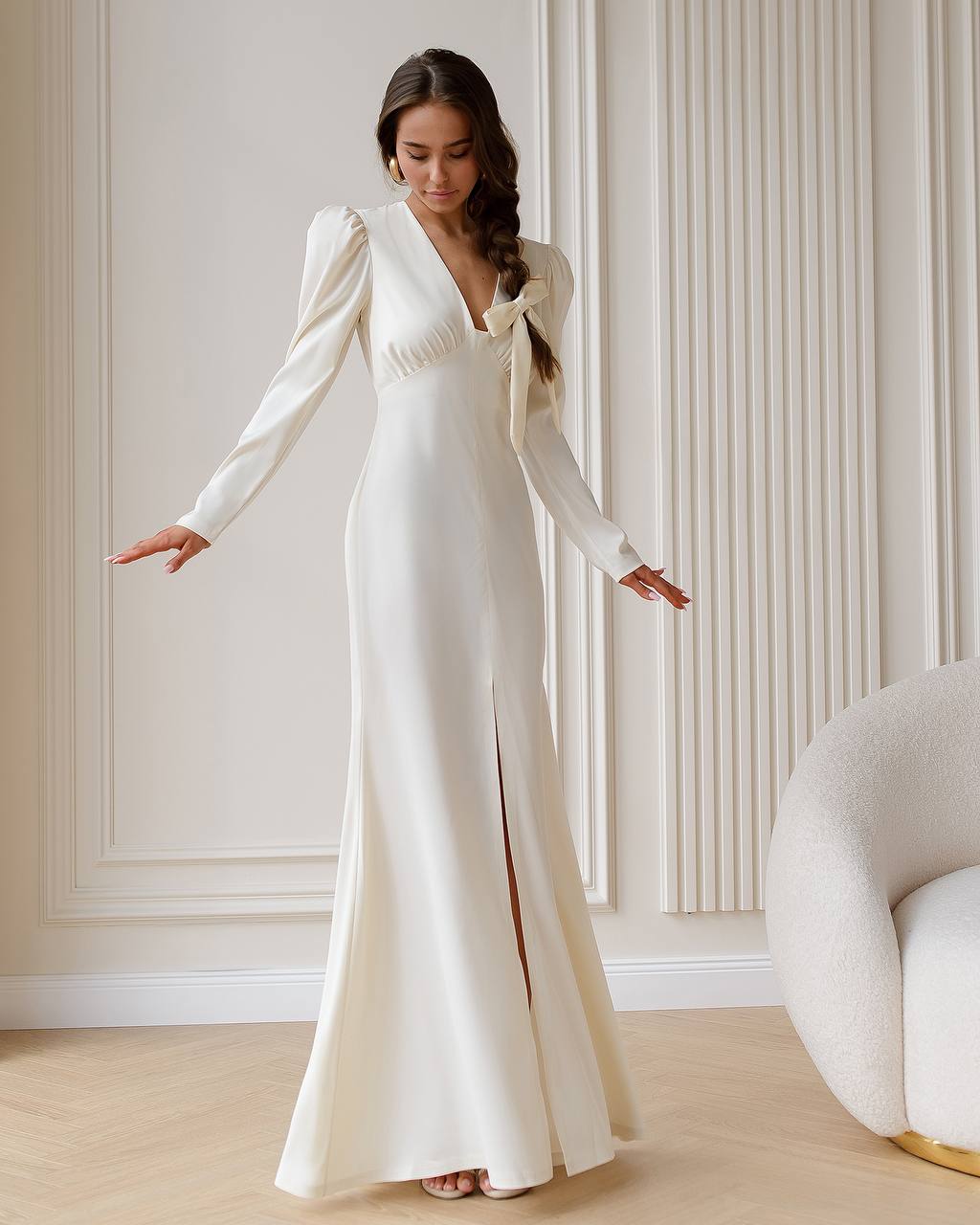 Woman in a white wedding dress standing in a room with light-colored walls and a chair.