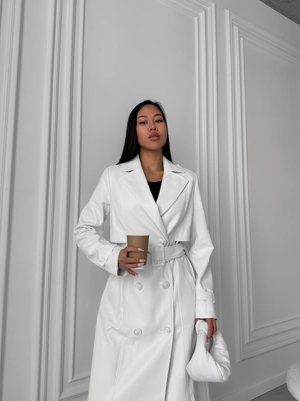 Woman in a white trench coat holding a coffee cup against a gray wall.