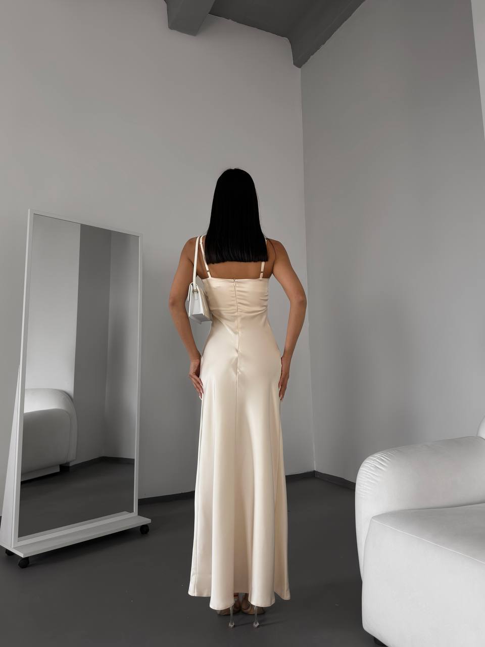 Woman in a beige evening gown standing in a minimalistic room with a mirror and white sofa.