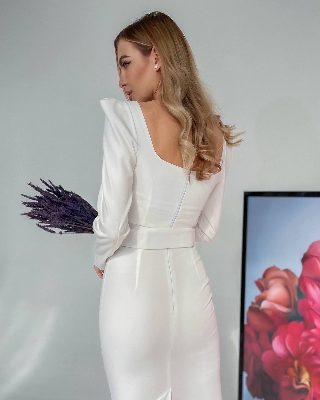 Woman in a white dress holding lavender against a neutral background with a colorful floral arrangement.