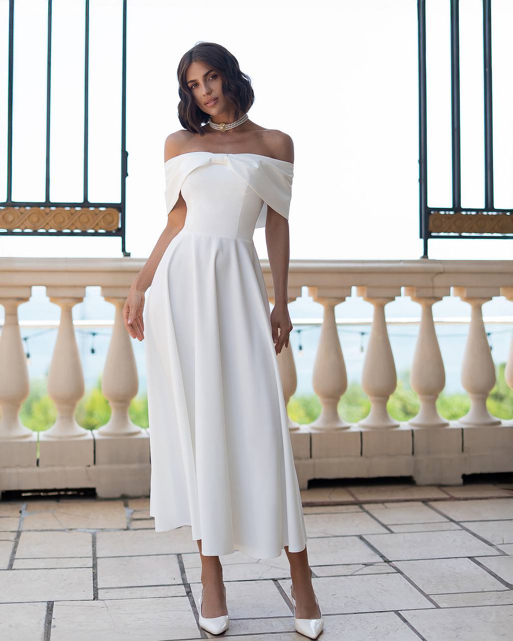Woman wearing a white off-shoulder dress on a balcony with a scenic background