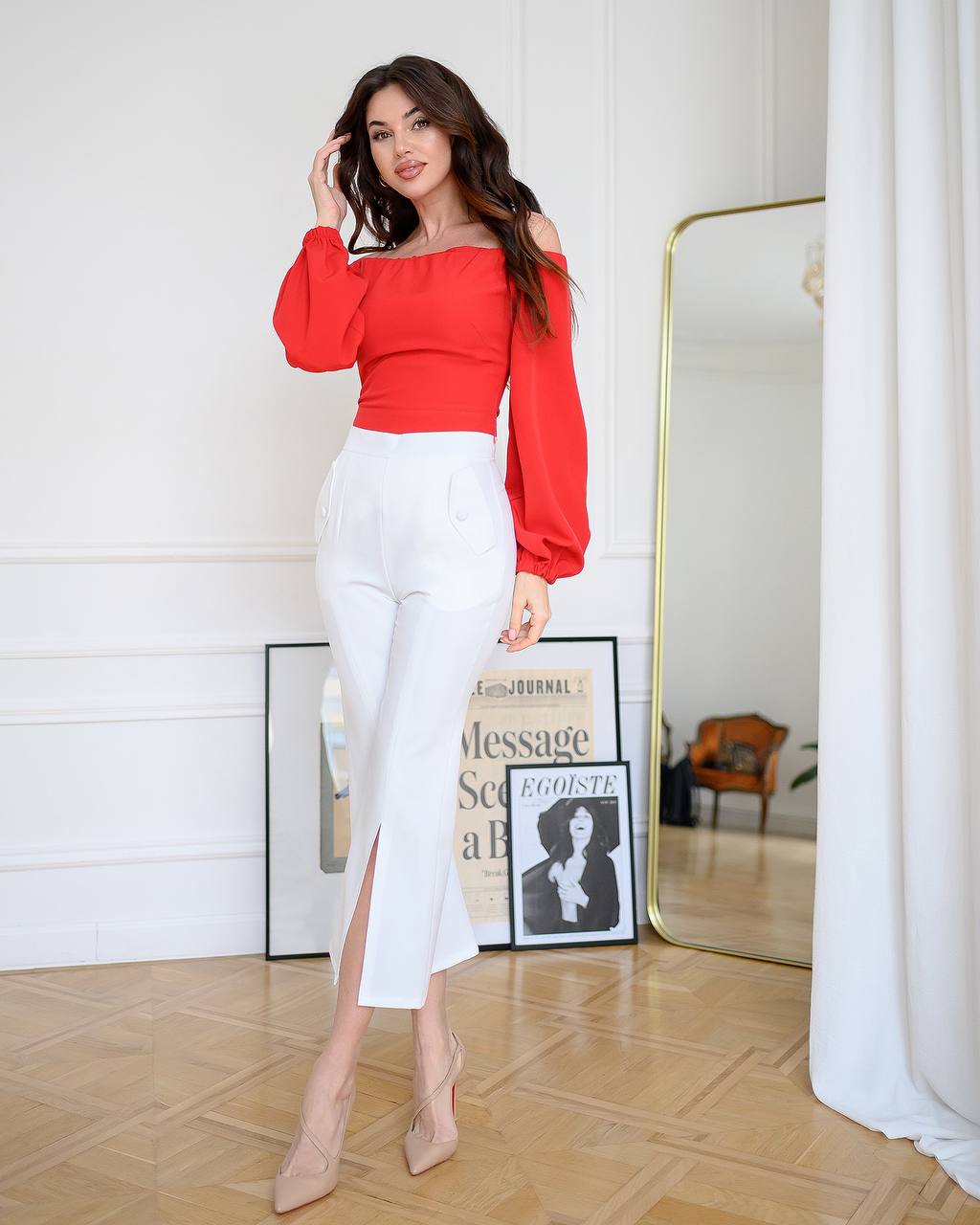 Woman in a red top and white pants standing in a stylish room with a mirror and decor.
