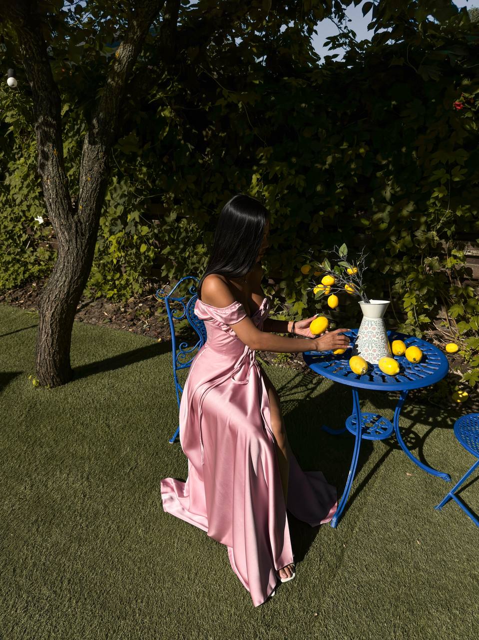 Woman in a pink dress standing next to a table with lemons and a vase outdoors.