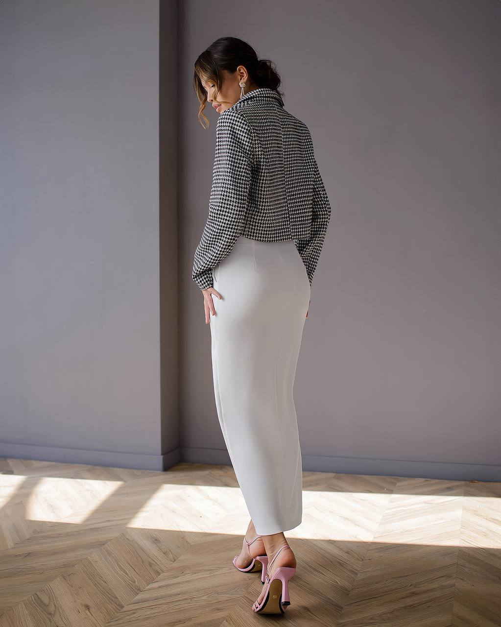 Woman in a checkered shirt and white pants standing in a room with a gray wall and wooden floor.