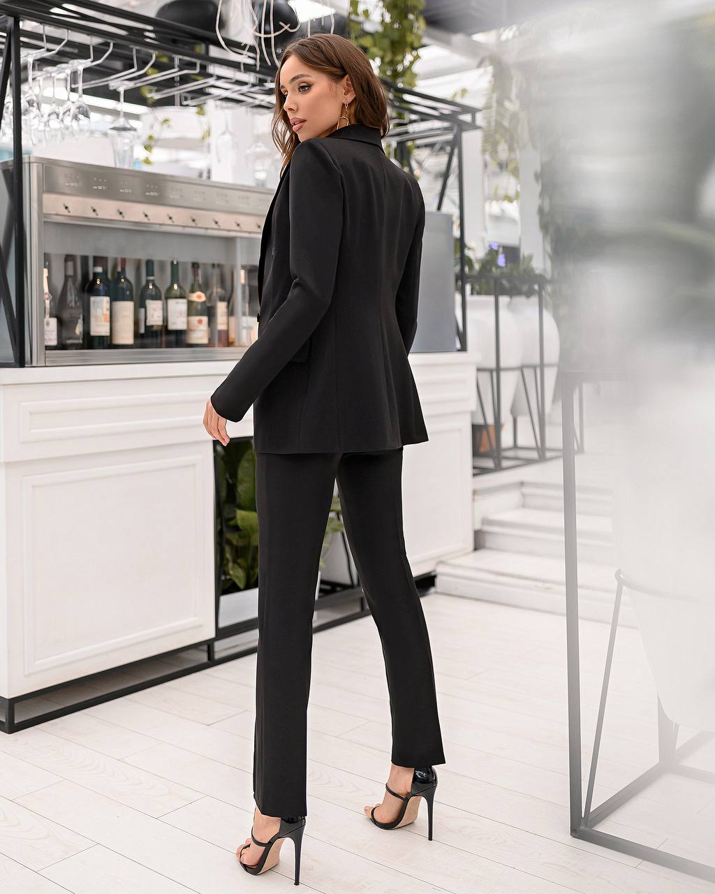 Woman in a black suit standing in a modern indoor setting with a bar in the background.
