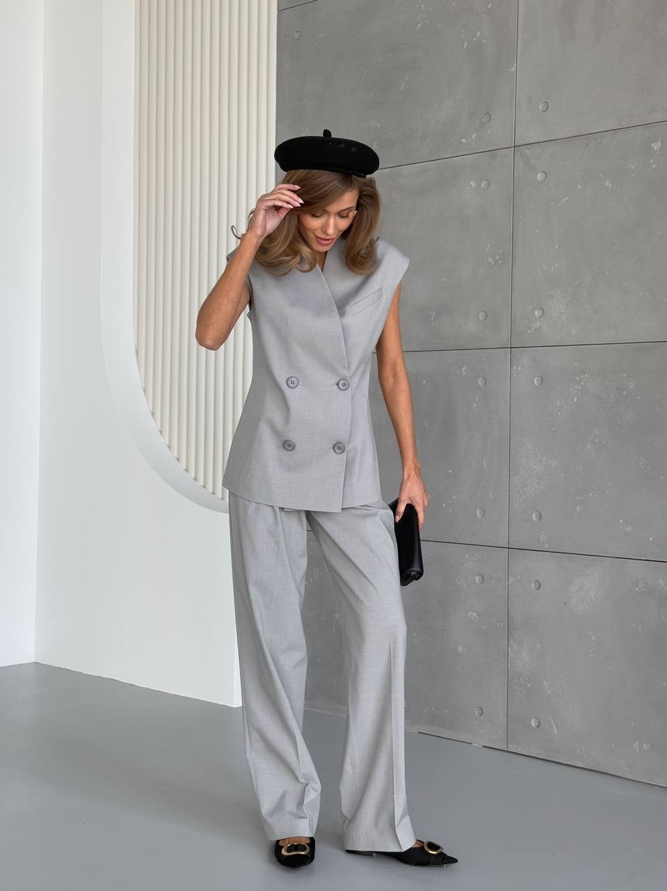Woman in a gray suit and black beret standing against a gray wall.