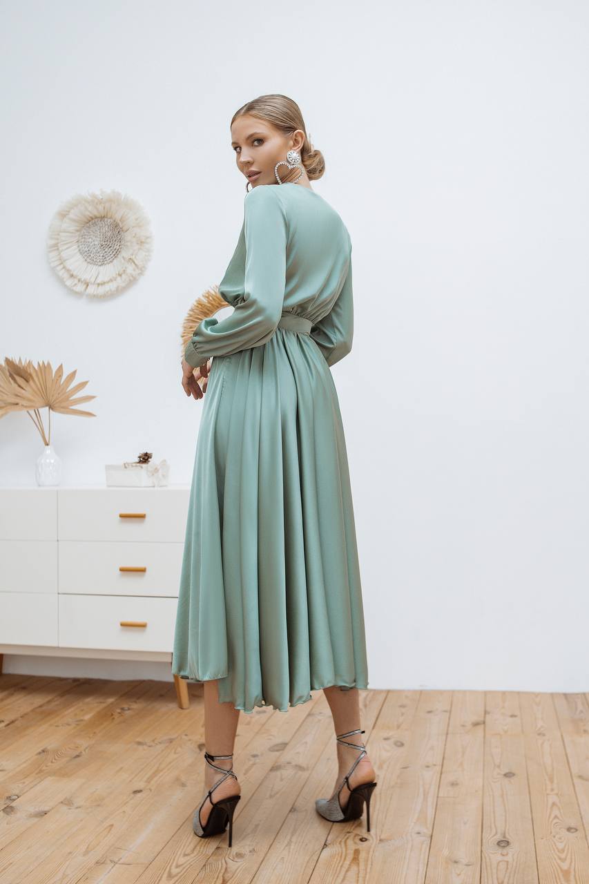 Woman wearing a light green dress standing in a room with wooden flooring and white walls.