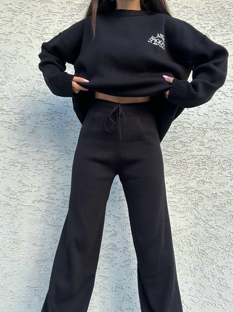 Person wearing a black oversized sweatshirt and pants against a textured wall.