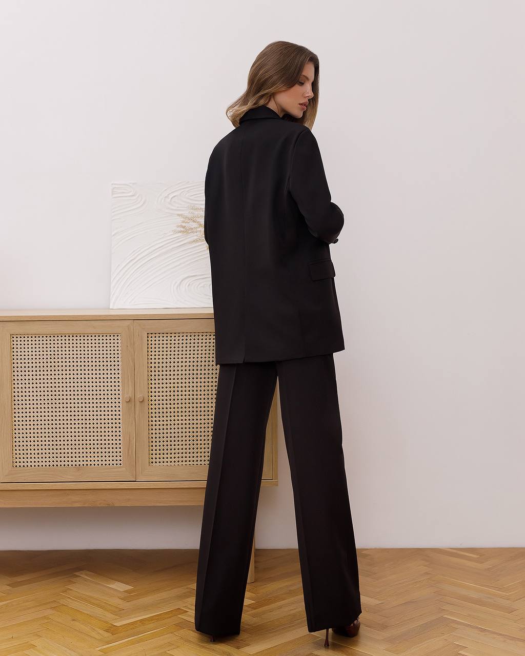Woman wearing a black suit standing in a room with wooden flooring and a light-colored wall.