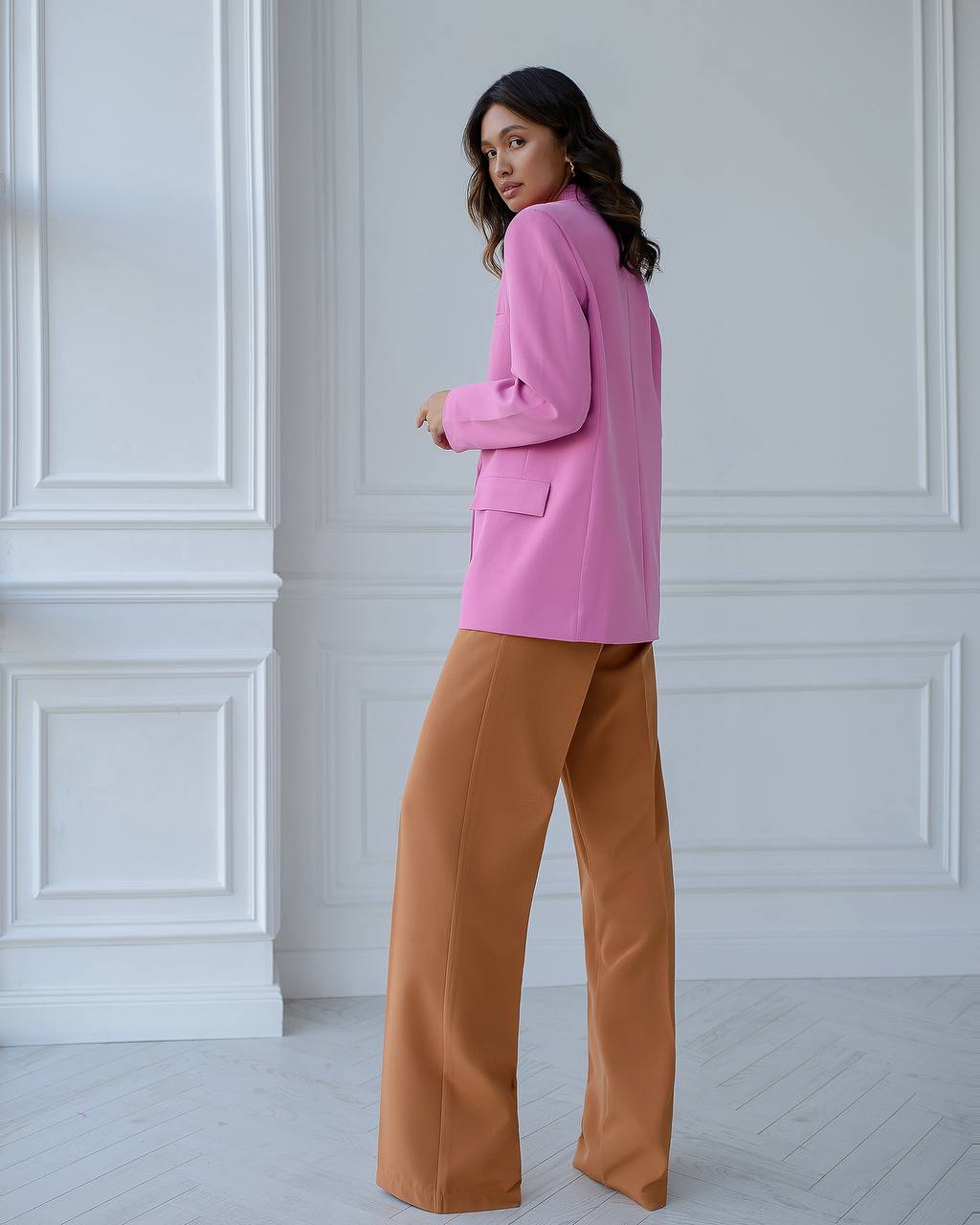 Woman wearing a pink blazer and brown pants against a white paneled wall.