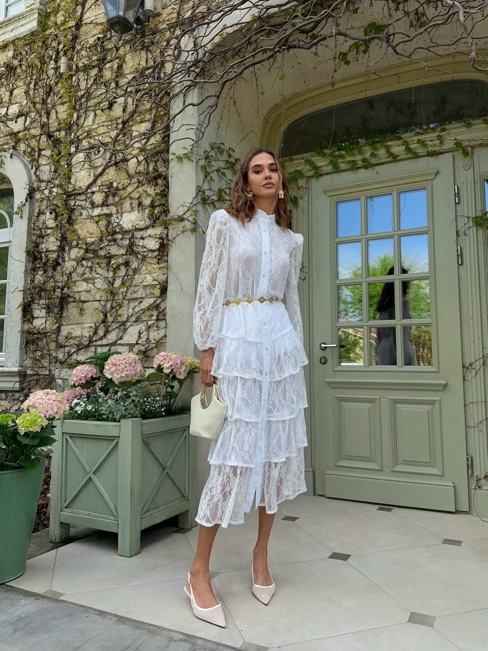 Woman in a white lace dress standing in front of a green door with floral decorations.