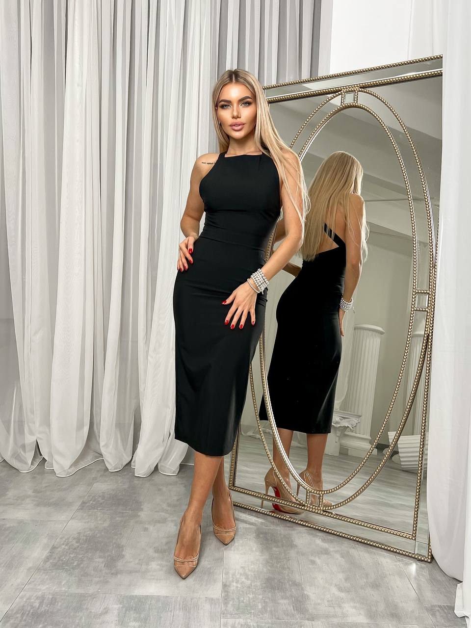 Woman in a black dress standing in front of a mirror in a room with white curtains.