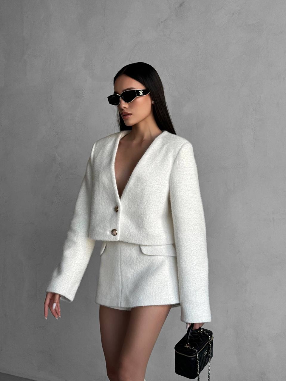 Woman in a white outfit with sunglasses and a black handbag against a gray background