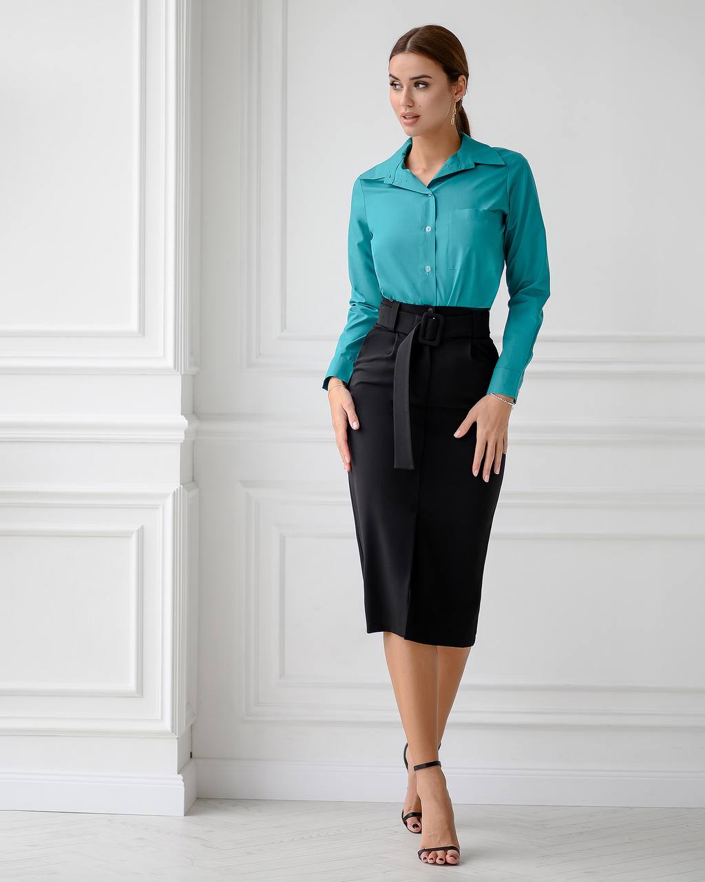 Woman wearing a teal shirt and black skirt against a white paneled wall.