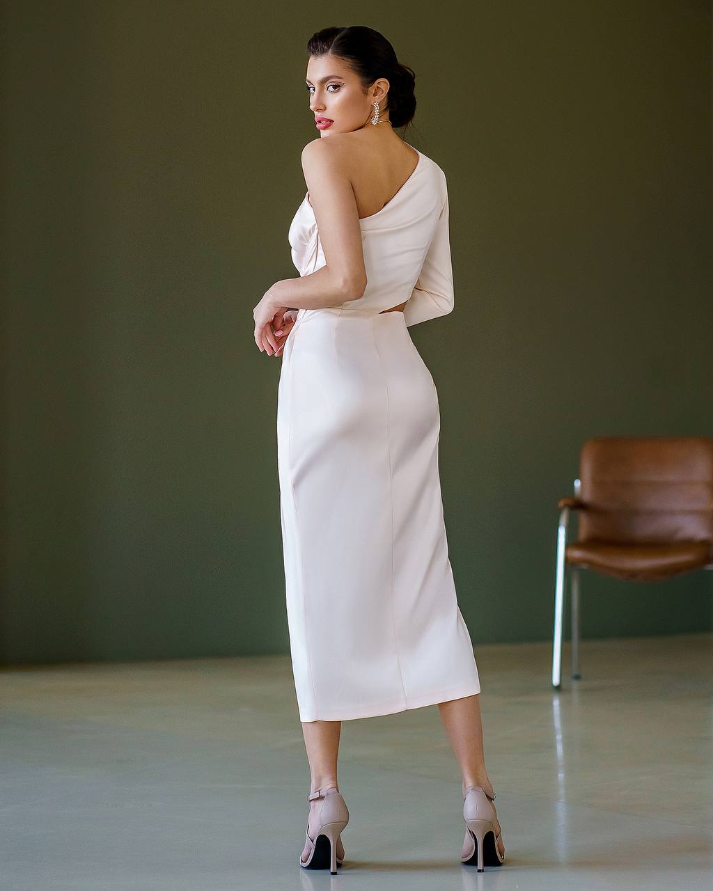Woman in a white one-shoulder dress standing in a room with a brown chair.