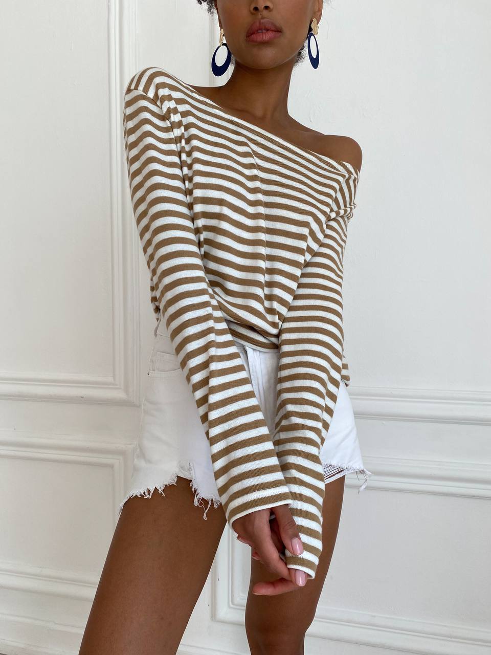 Woman wearing a striped off-shoulder top and white shorts against a white wall.