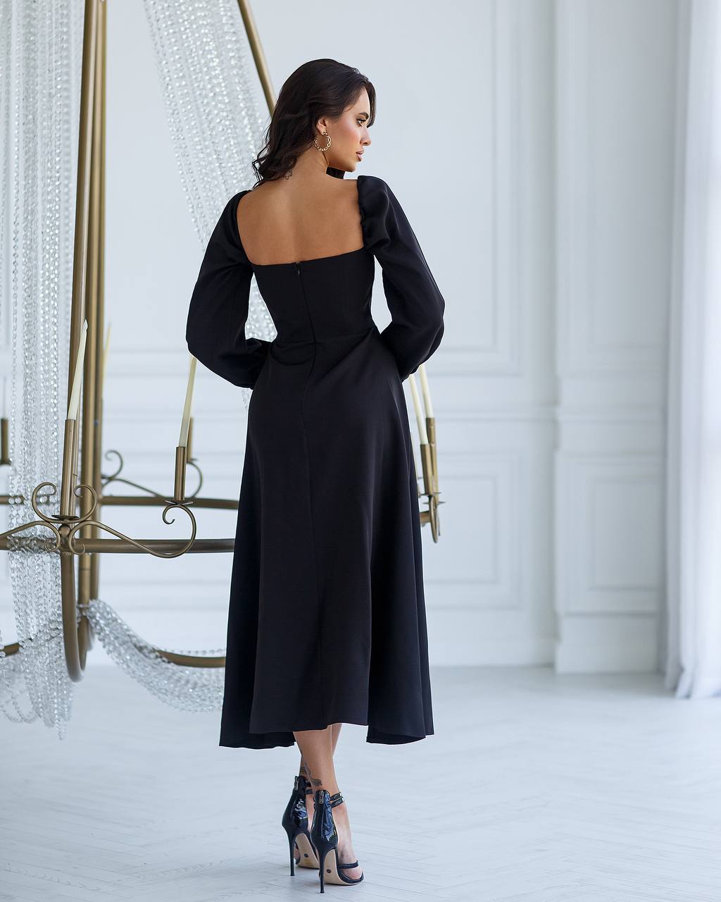 Woman wearing a black dress with long sleeves and open back in a white room.