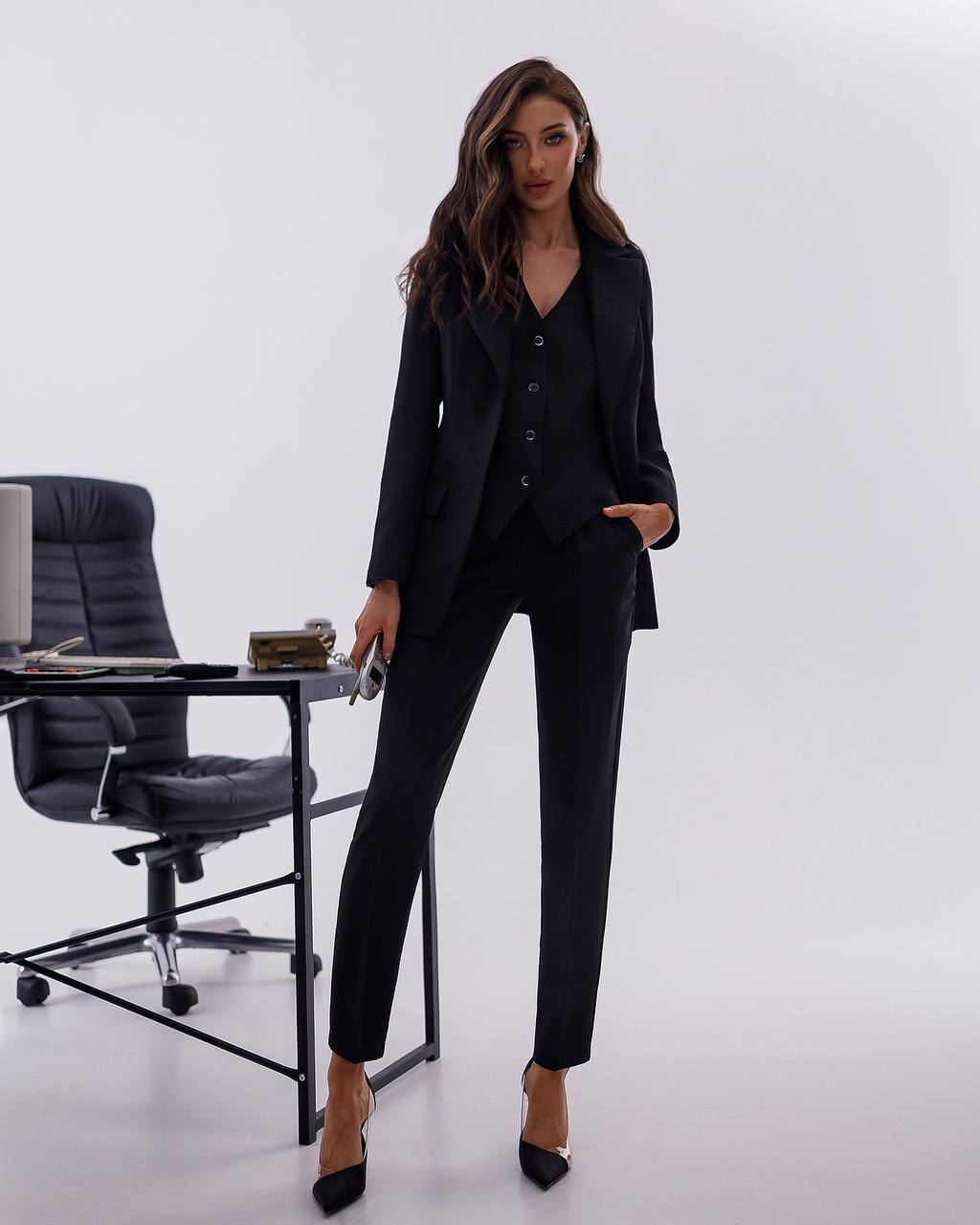 Woman in a black suit standing in an office setting with a chair and desk.