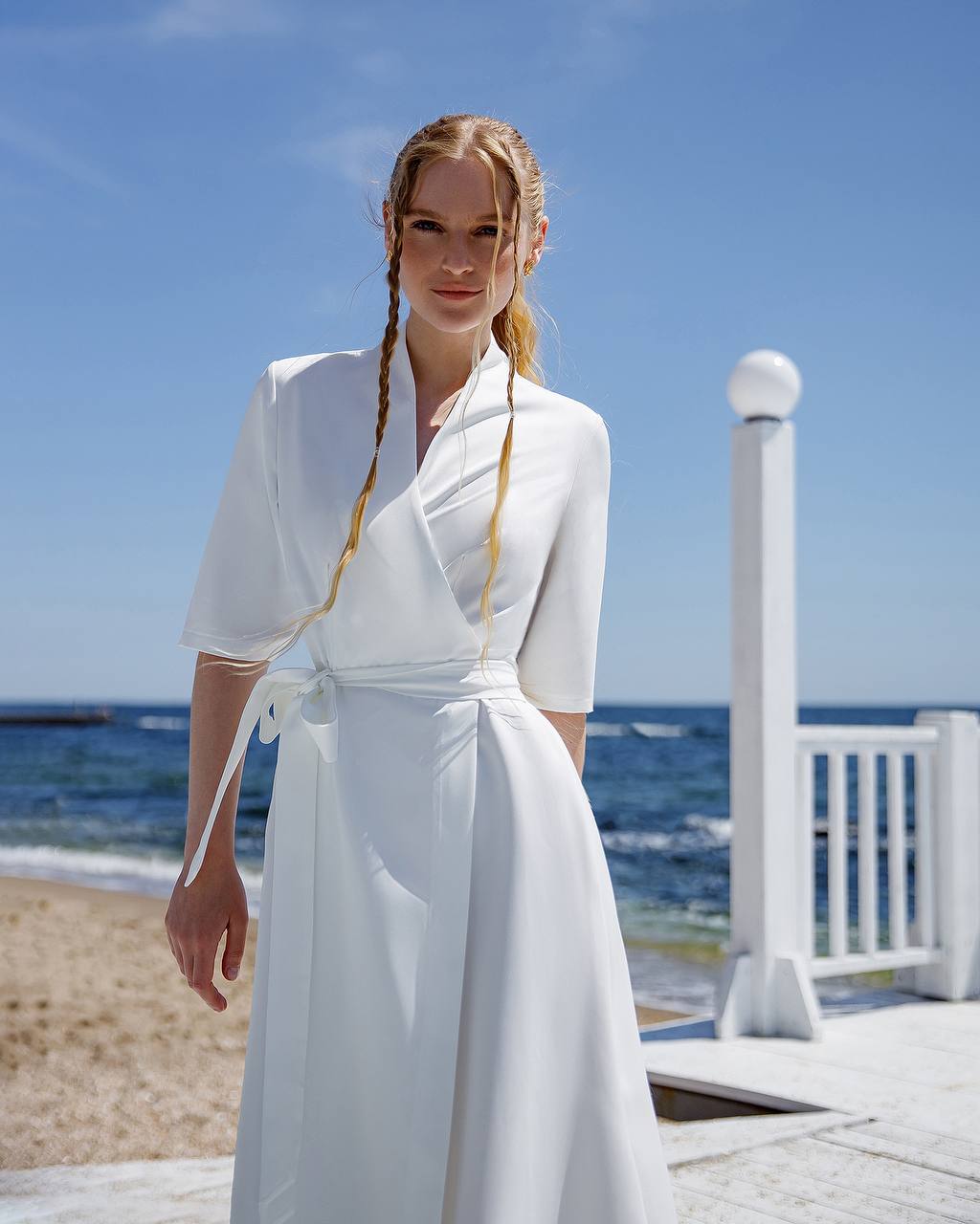 Woman in a white dress standing on a beach with clear blue sky and ocean.