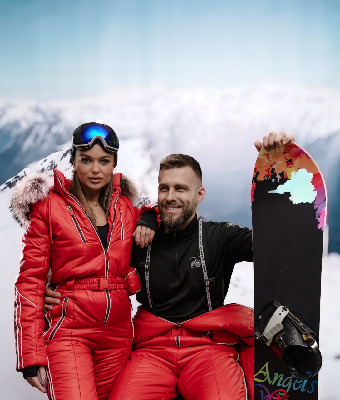 Two people in ski gear with a snowboard against a mountainous background