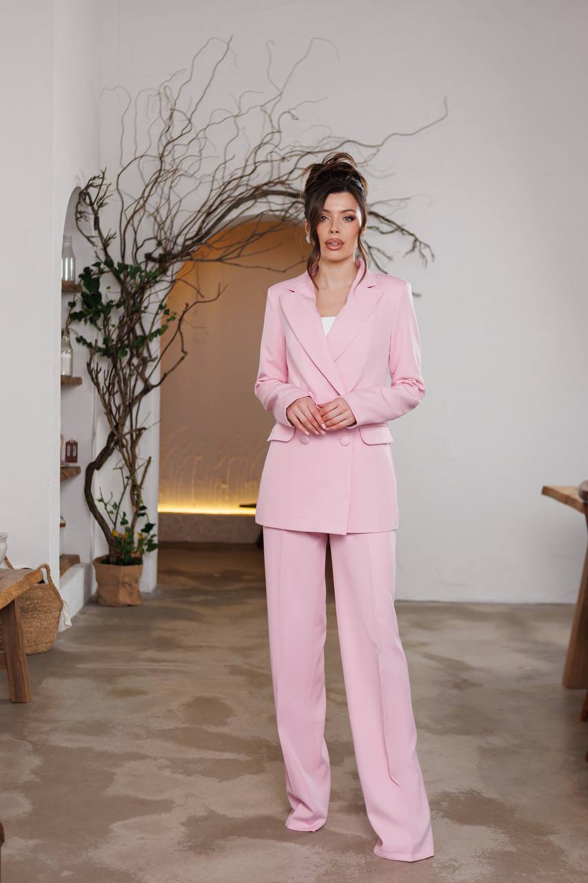 Woman in a pink suit standing in an indoor setting with plants and a neutral wall.