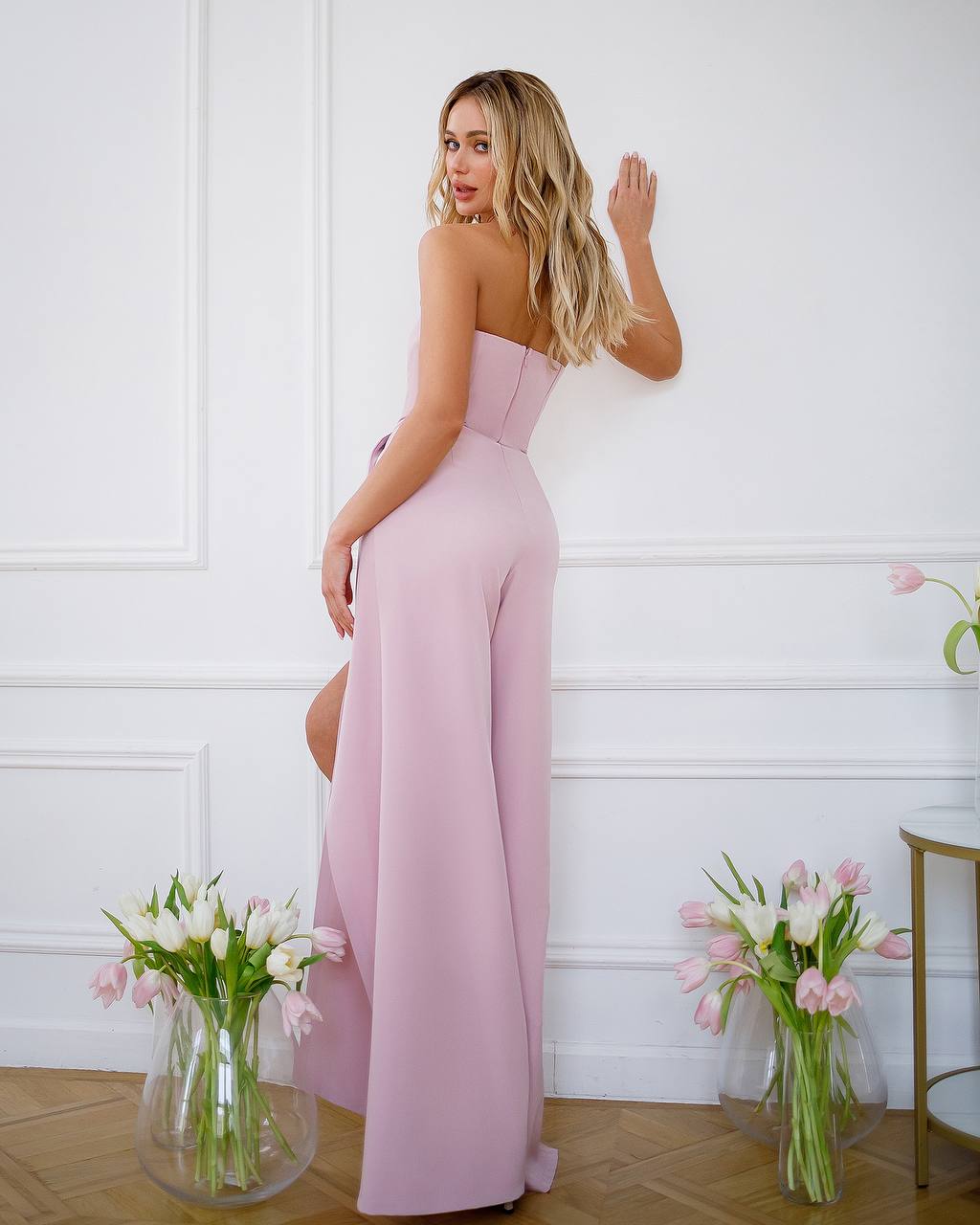 Woman wearing a pink strapless jumpsuit in a room with white walls and floral decorations.