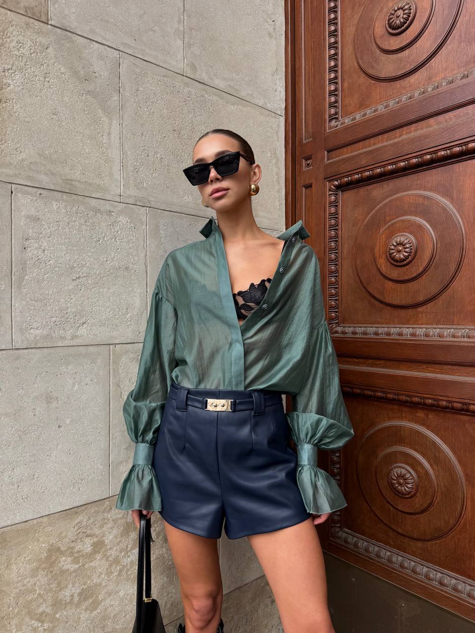 Woman in green shirt and blue shorts standing against a textured wall with a wooden door.