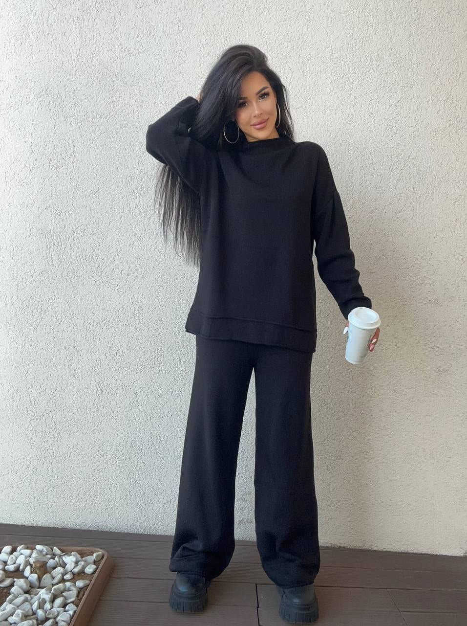 Woman wearing a black outfit holding a white cup against a plain wall.