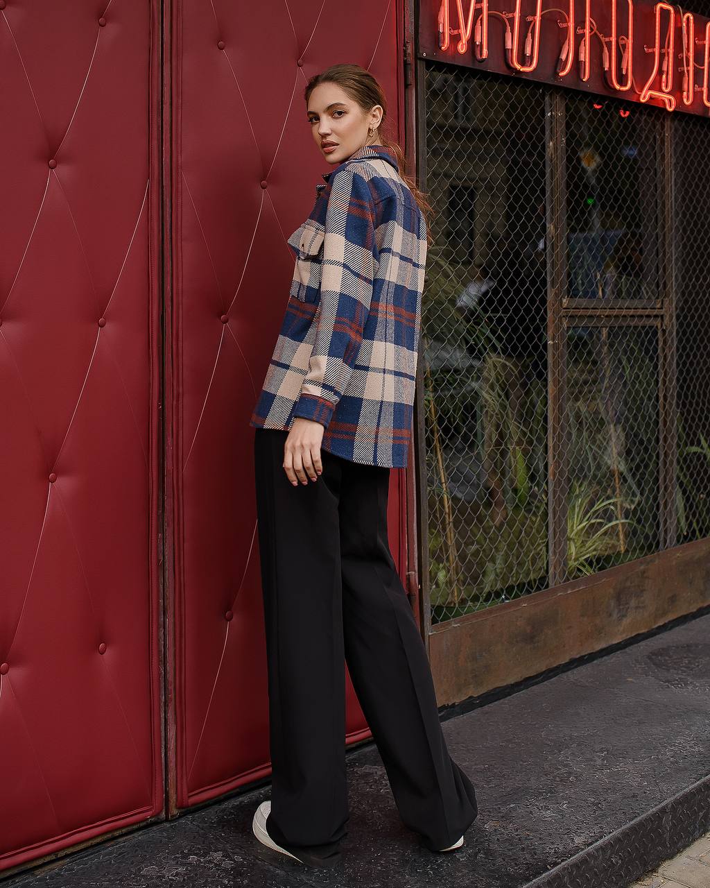 Woman in a plaid shirt and black pants standing against a red wall with neon signs.