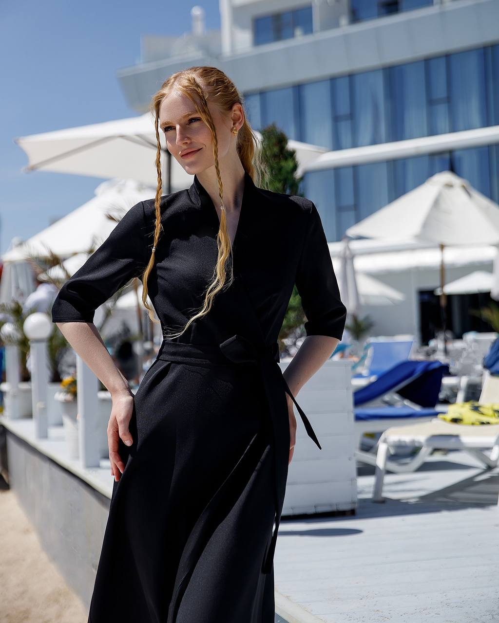 Woman in a black dress standing on a beach with umbrellas and lounge chairs in the background.